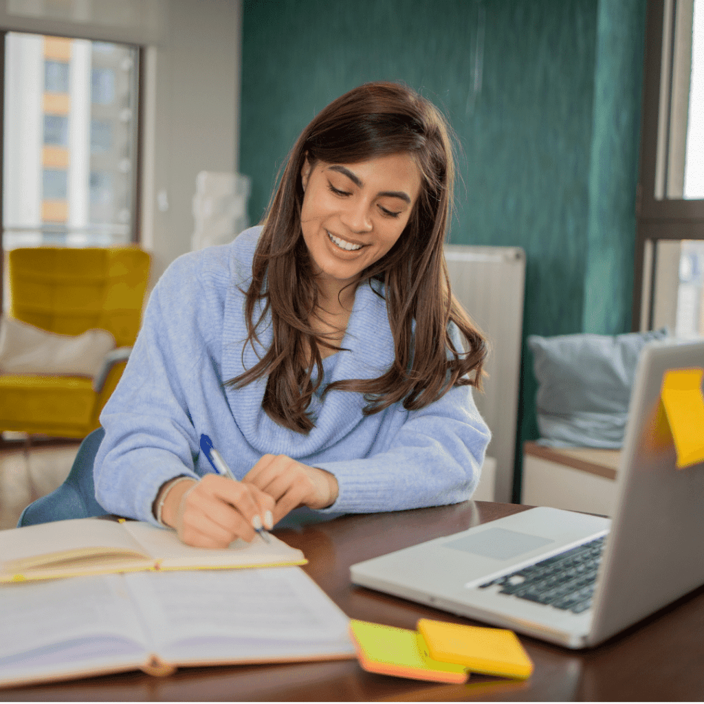 Frau arbeitet lächelnd mit Laptop und Notizbuch.